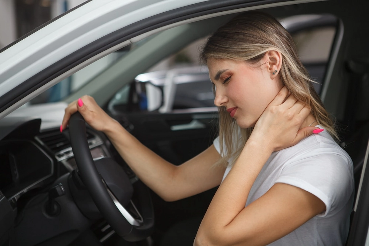 A woman sitting in the driver’s seat of a car holds her neck in pain, suggesting discomfort or possible whiplash after an incident.