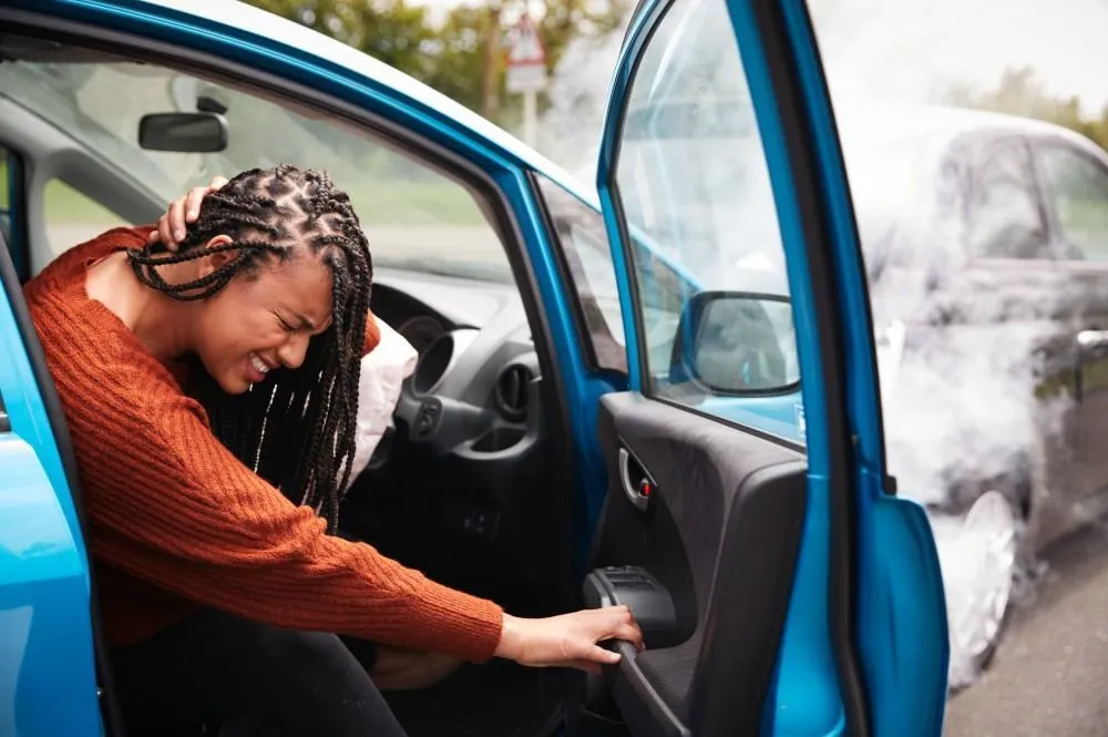 A young Black woman with braided hair sits in a blue car after a collision, holding her head in pain, with a deployed airbag and smoke coming from the damaged car outside.
