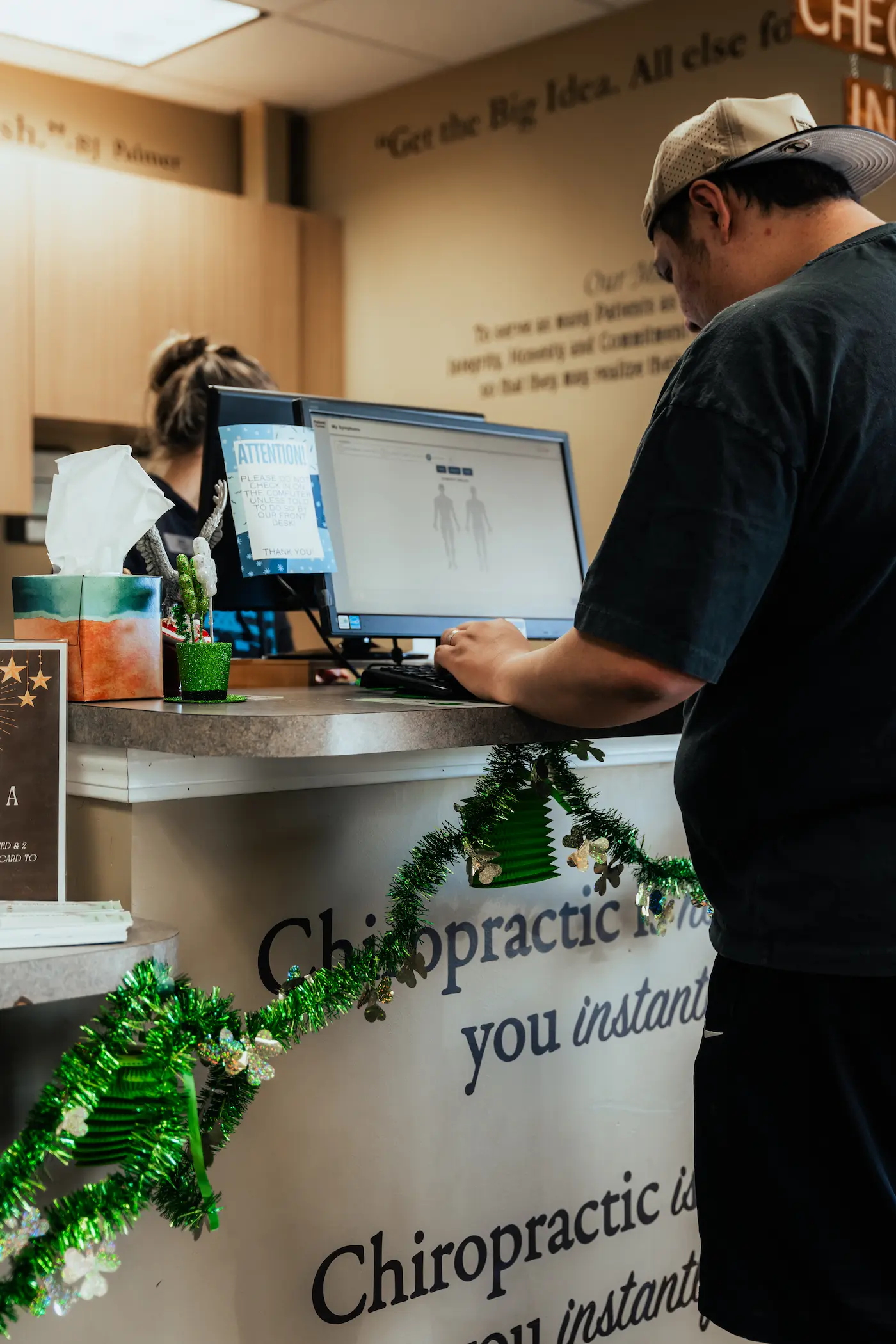 A man in a baseball cap uses a computer at a reception desk decorated with green tinsel and shamrocks, with a receptionist visible in the background.