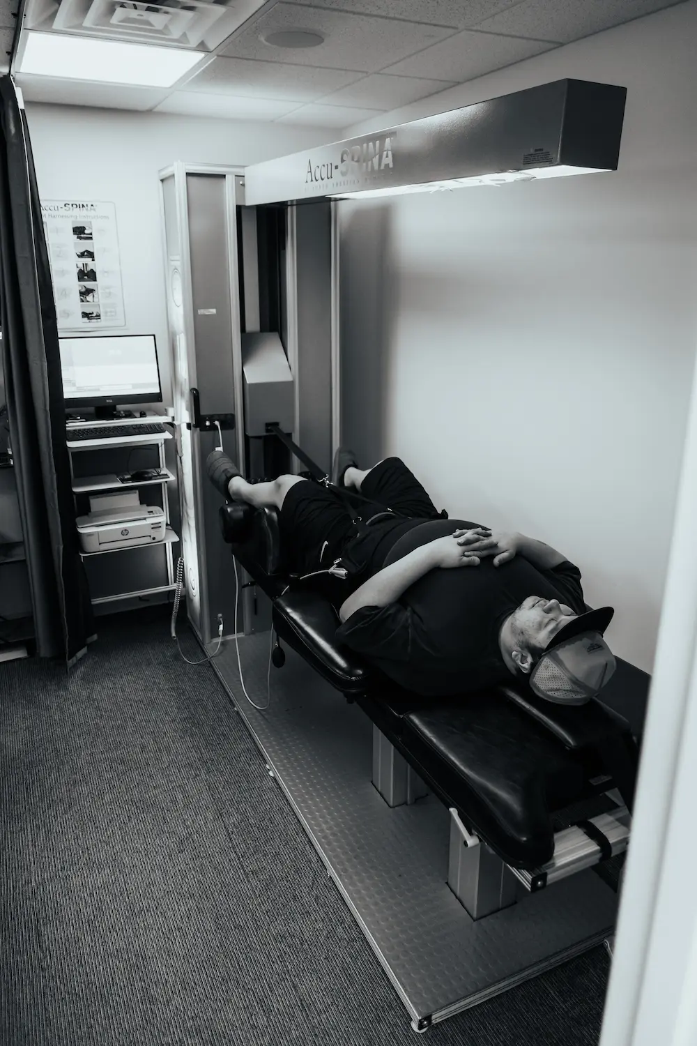 Black and white image of a man lying on a treatment table, connected to an Accu-SPINA spinal decompression machine for therapy.