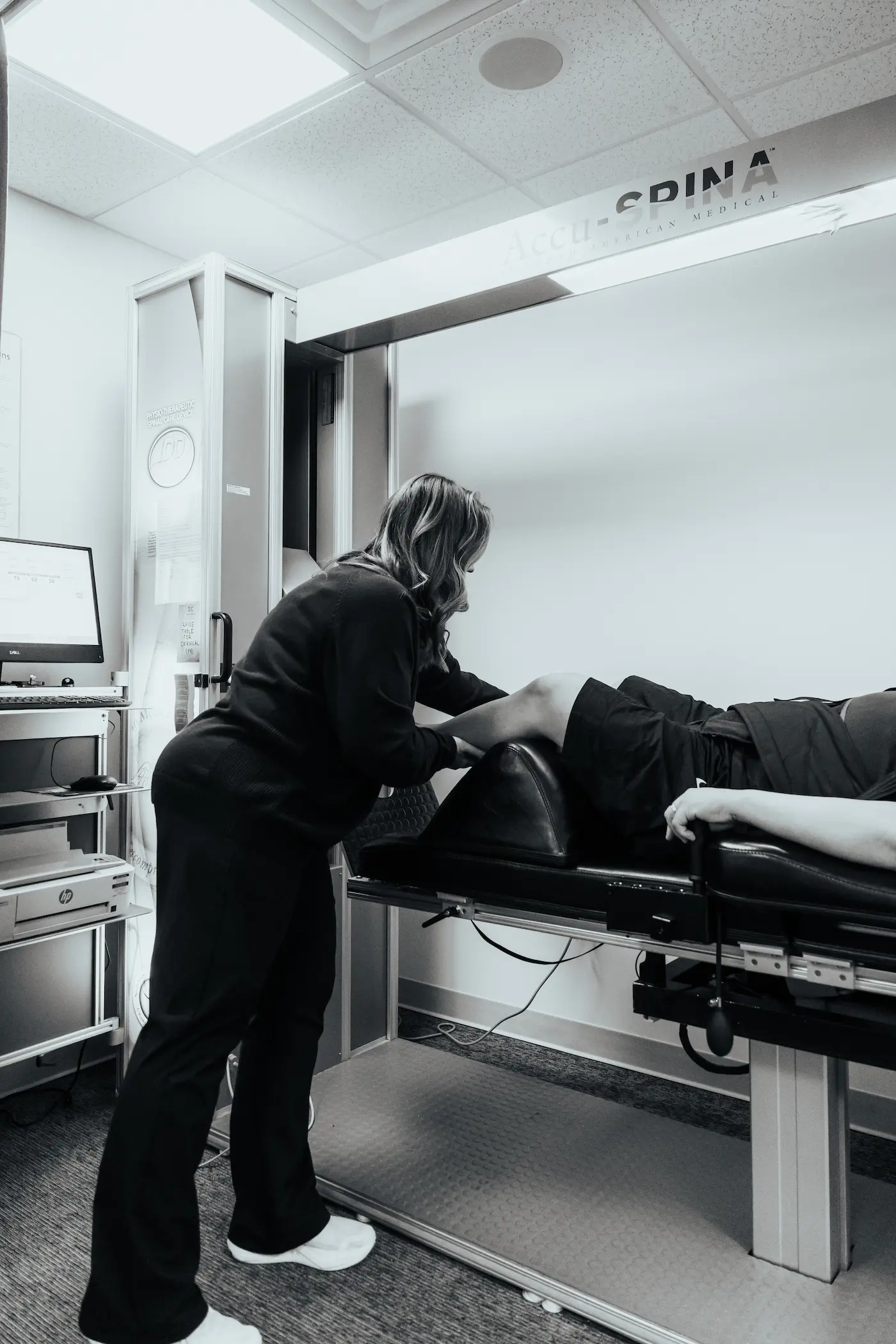 A healthcare professional in dark scrubs assists a patient lying on an Accu-SPINA medical device in a monochrome clinical photo.