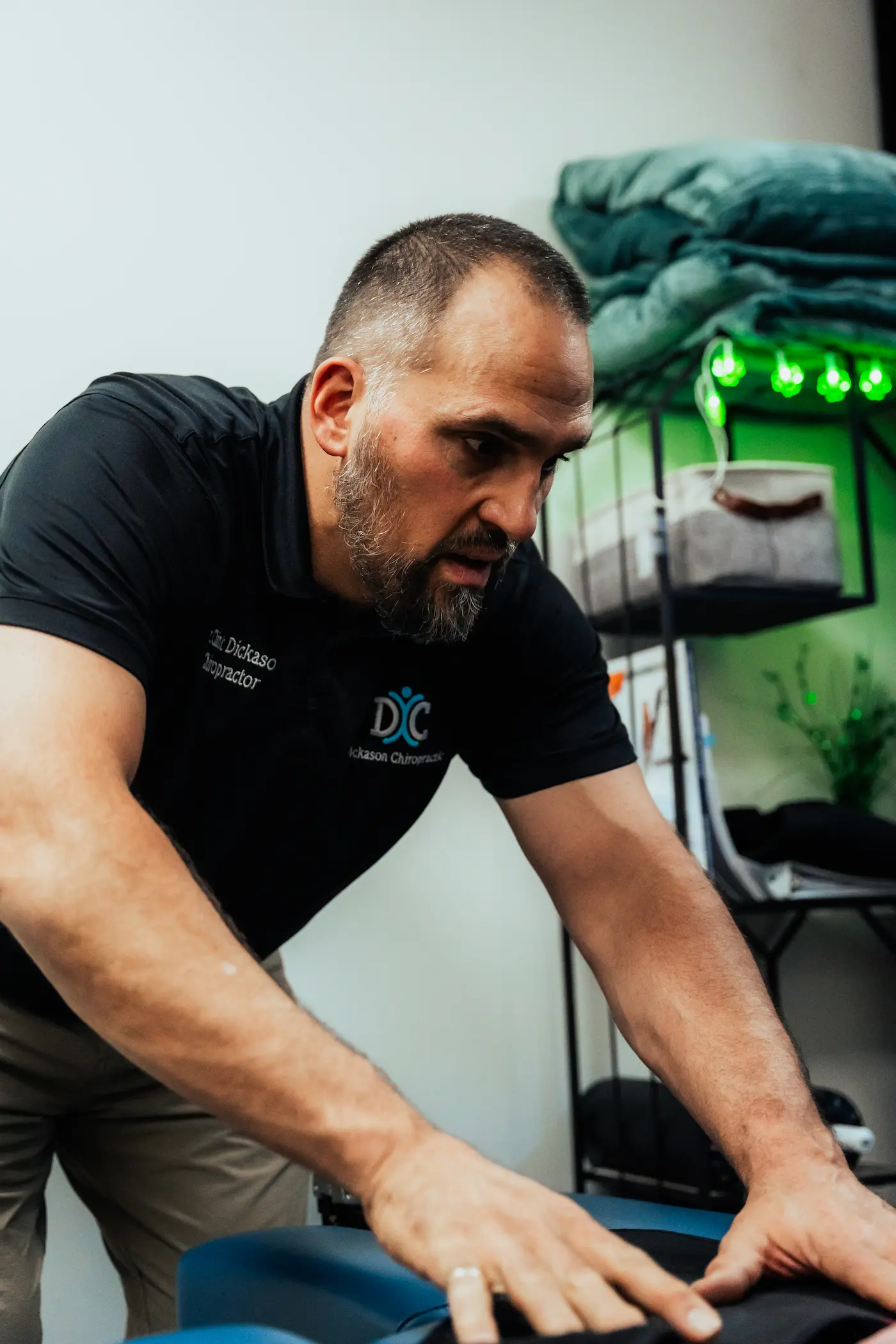 A male chiropractor, Dr. Dickason, wearing a black polo shirt, leans over a blue treatment table, placing his hands on a patient's back with focused concentration. Green lights are visible in the blurred background.