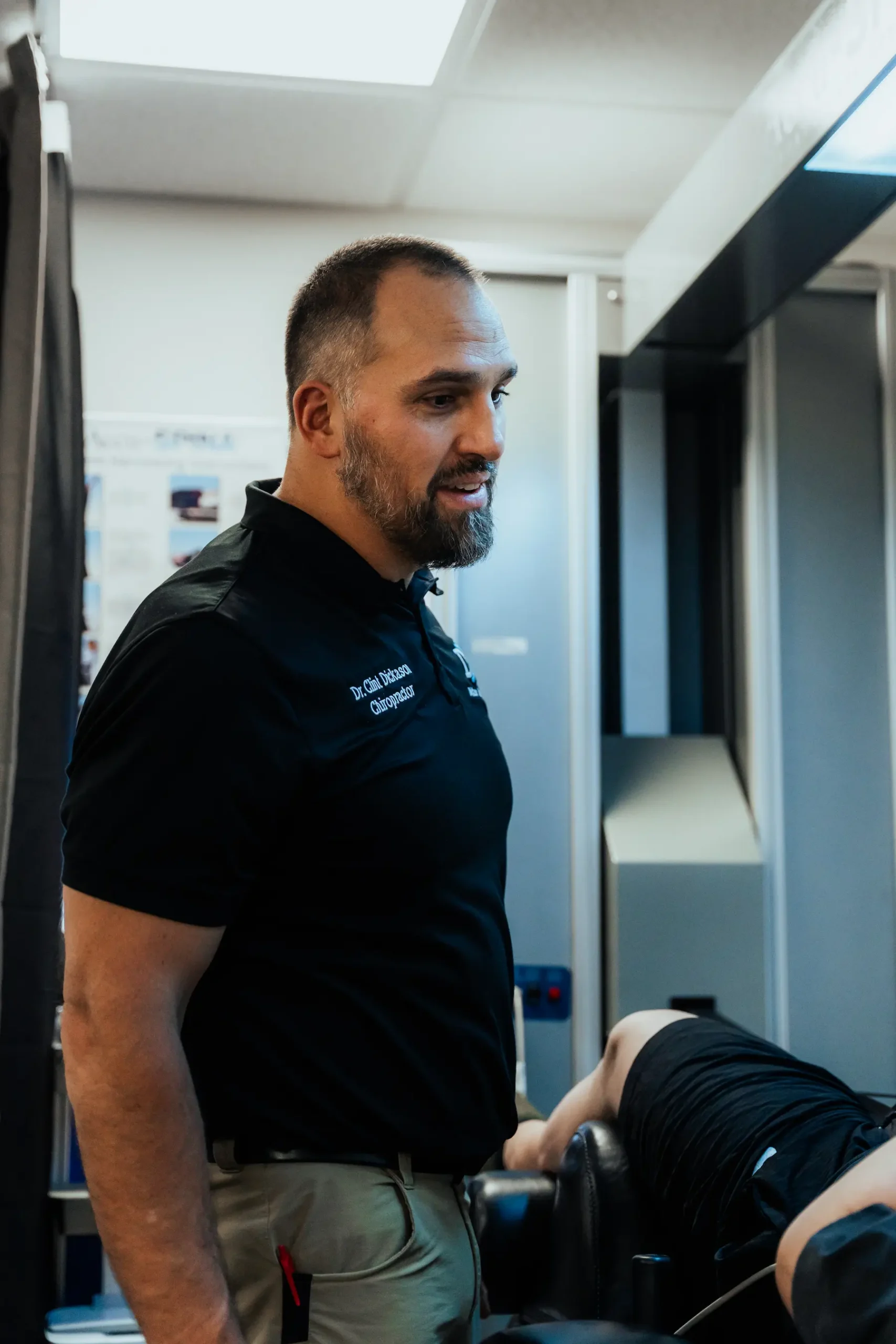 A male chiropractor with a beard, wearing a black polo shirt, looks intently at a patient's legs on a black treatment table in a clinic.