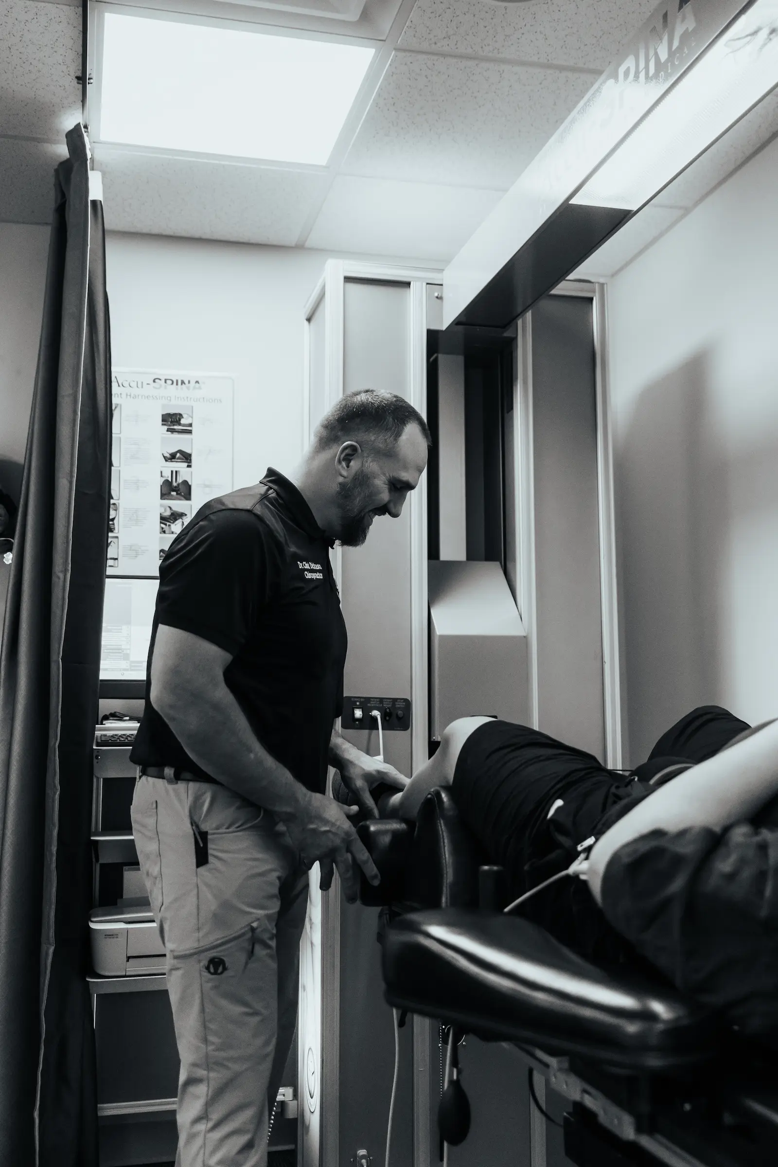 A male chiropractor with a beard, wearing a polo shirt, smiles down at a patient's leg on a treatment table in a black and white image. Medical equipment is in the background.
