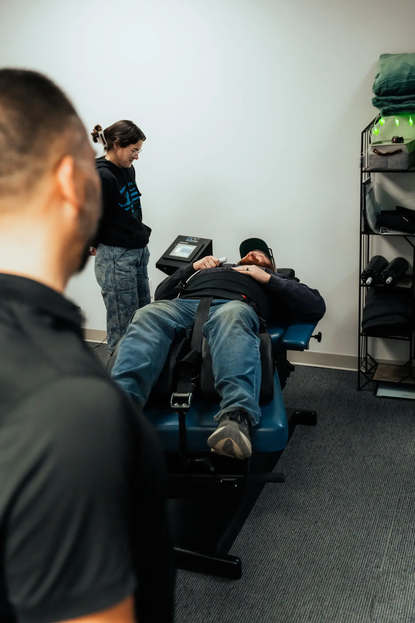 A man lies on a blue medical table, strapped in for therapy, while a woman stands nearby observing. Another person is in the foreground with their back to the camera. The room is a clean medical office.