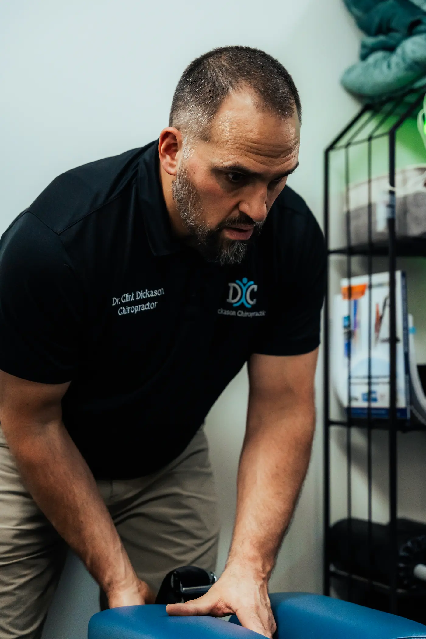 A male chiropractor, Dr. Clint Dickason, wearing a black polo shirt, leans over a blue adjustment table with focused intensity, his hands on the table.