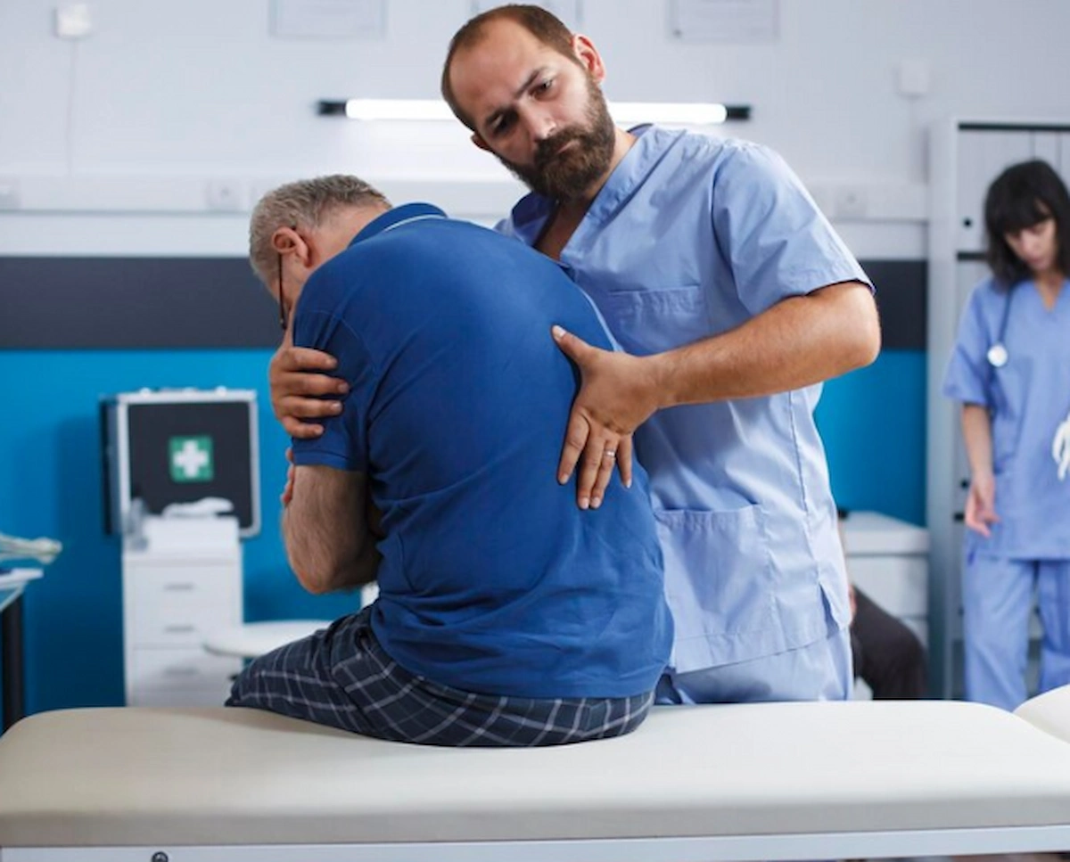 A male healthcare professional in blue scrubs performs a chiropractic adjustment on a seated male patient, placing his hands on the patient’s lower back while the patient is hunched over in a medical examination room.