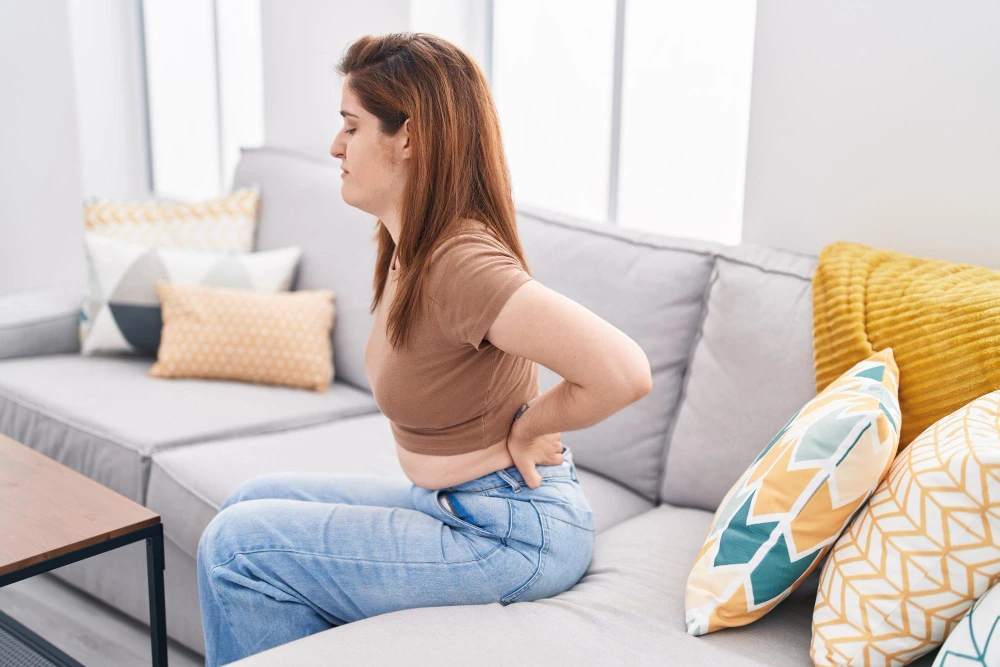Woman sitting on a sofa holding her lower back in pain, indicating discomfort in the lumbar area