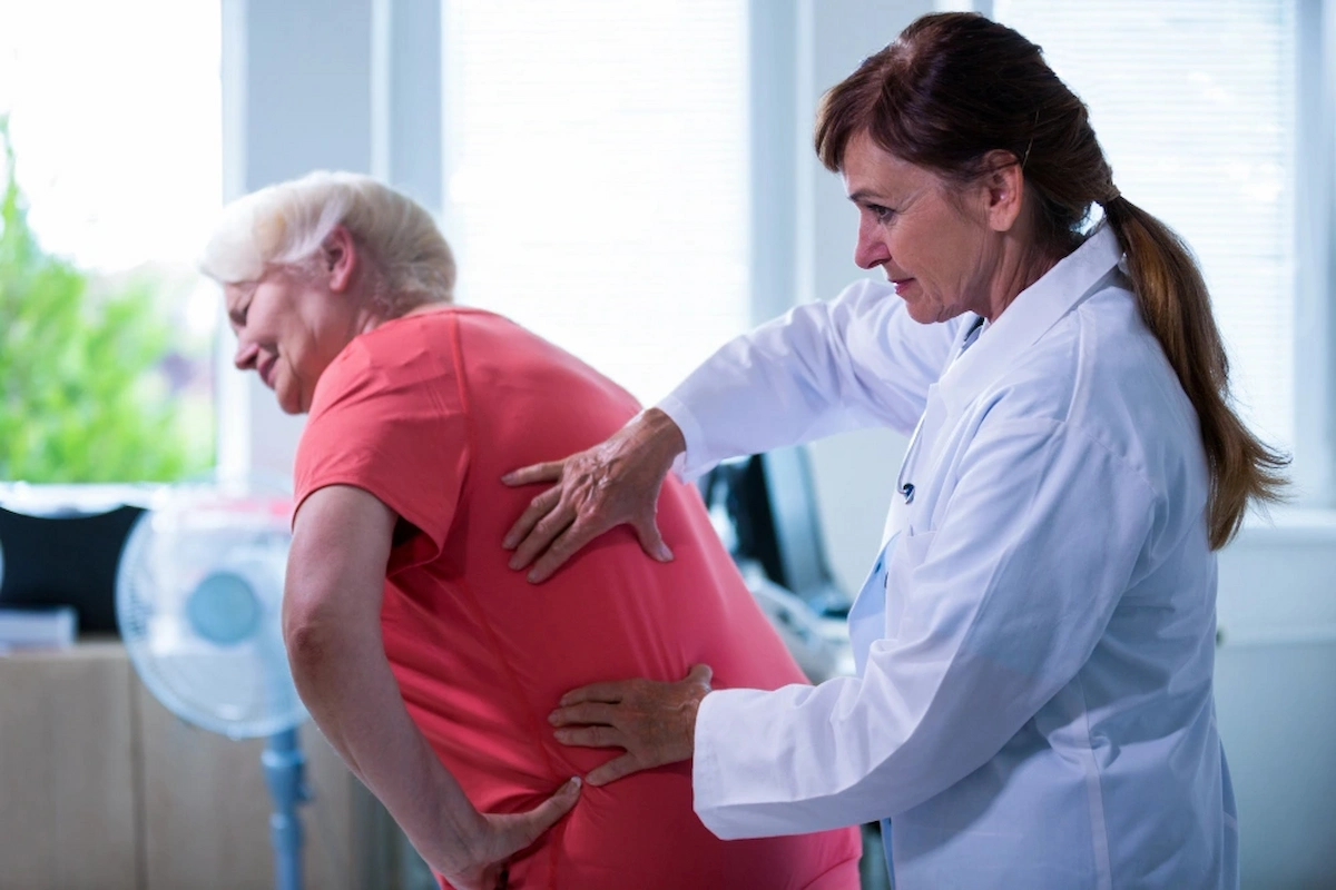 A doctor in a white lab coat assesses an older woman who is bent over, holding her lower back in pain, likely due to sciatica.