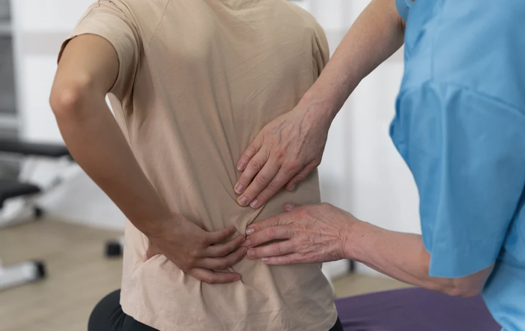 A medical professional in blue scrubs examines a patient’s lower back while the patient holds their back in discomfort, suggesting lower back pain or sciatica.