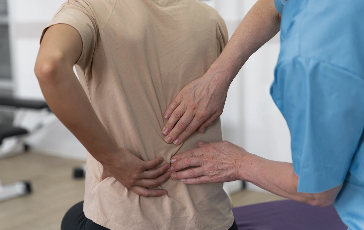 A medical professional in blue scrubs examines a patient’s lower back while the patient holds their back in discomfort, suggesting lower back pain or sciatica.