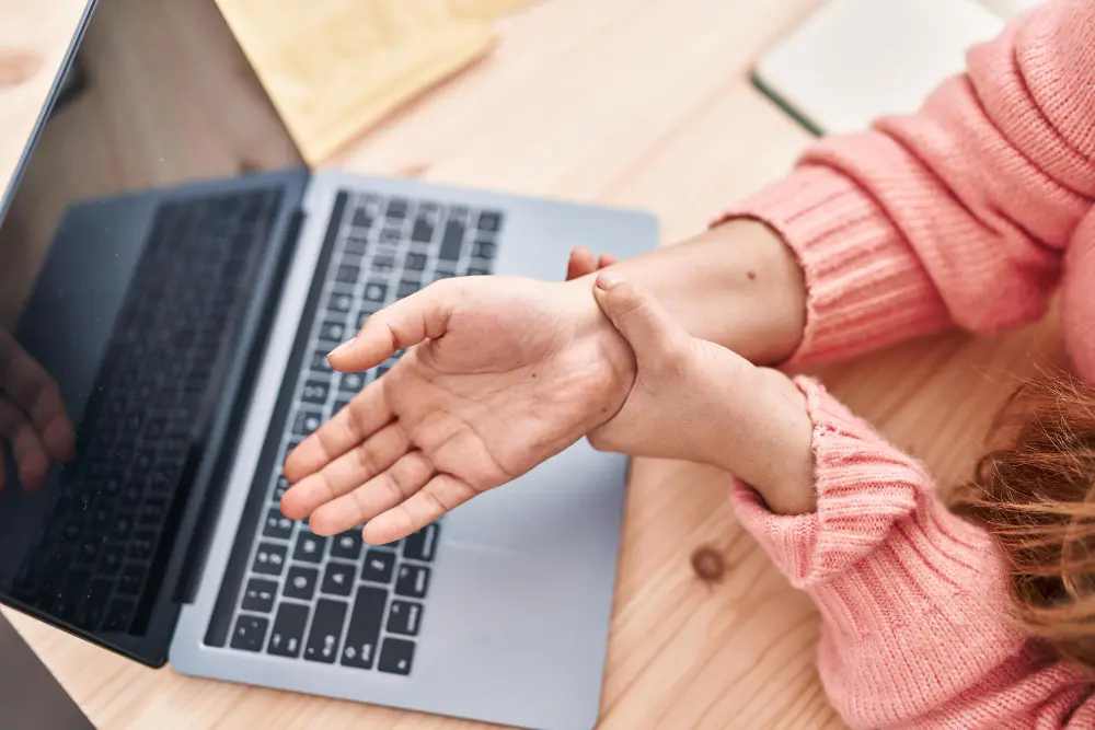 Overhead view of a person in a pink sweater holding their wrist in pain while sitting at a laptop on a wooden desk.