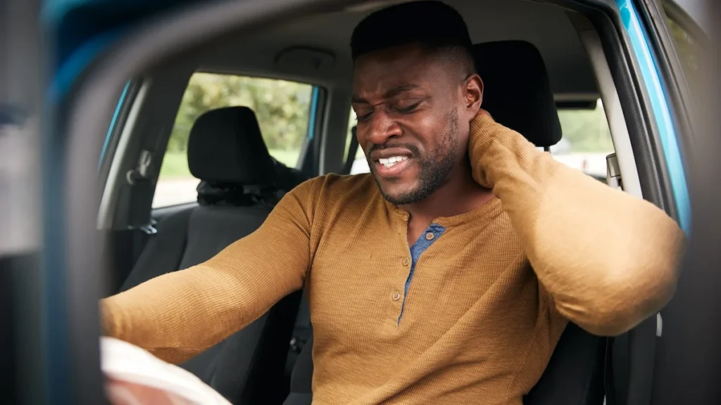 A man in a car, grimacing and holding his neck and shoulder in pain.