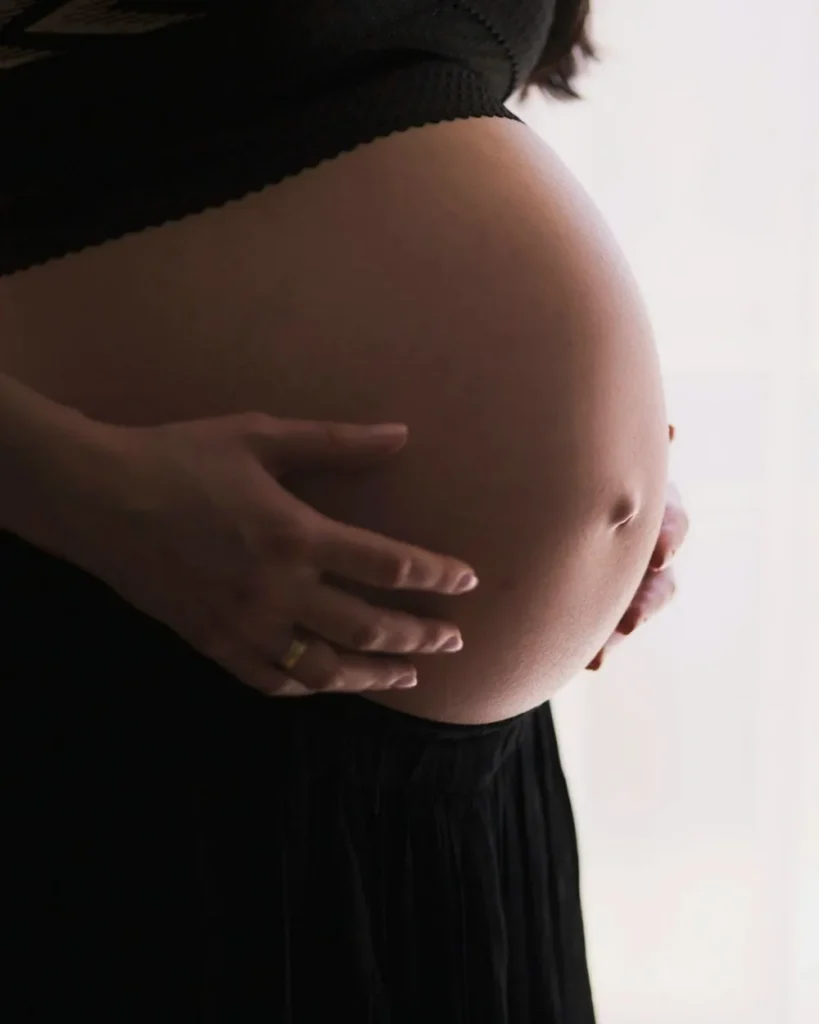 Close-up of a pregnant belly, with hands gently cradling it. A gold ring is visible on one hand. The background is bright white.