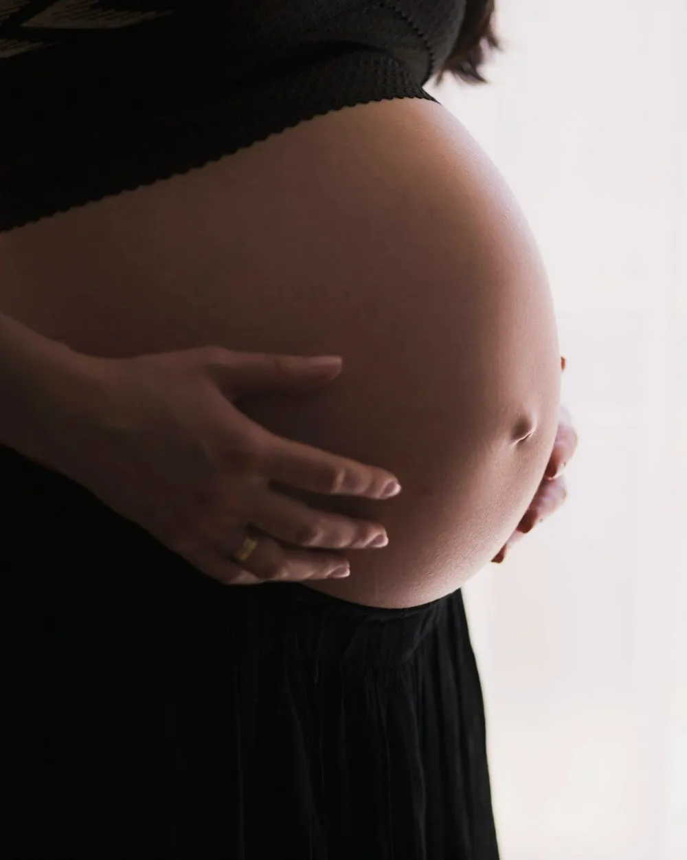 Close-up of a pregnant belly, with hands gently cradling it. A gold ring is visible on one hand. The background is bright white.