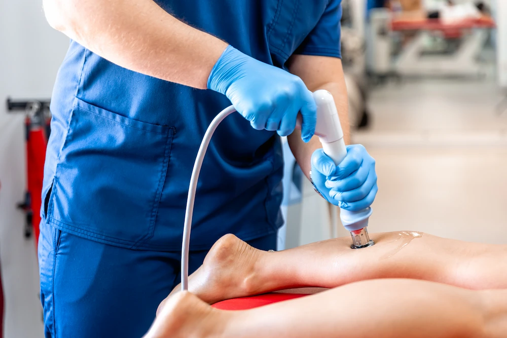 A person in blue scrubs and gloves uses a white medical device on a patient's leg, which has clear gel applied.